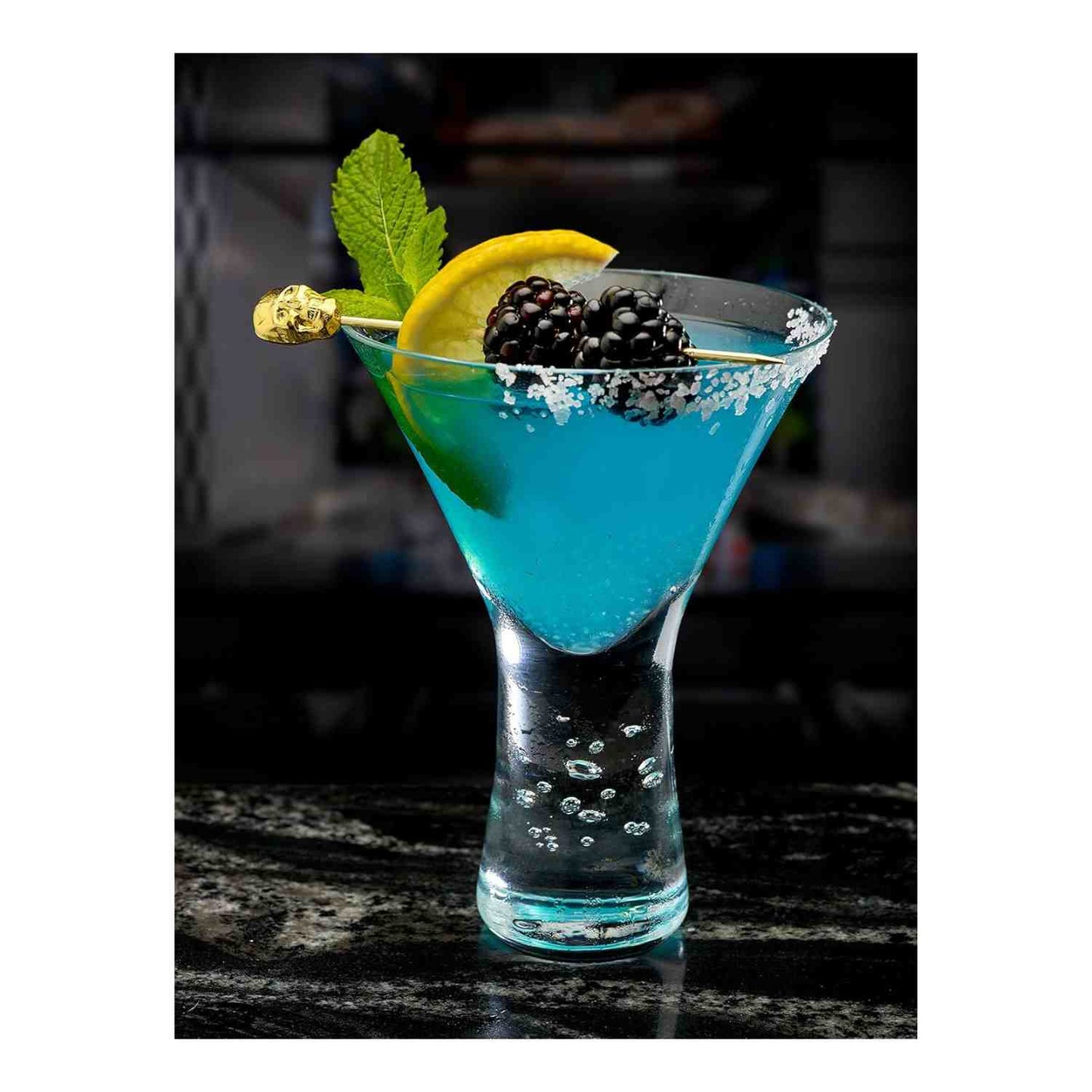 A bright blue cocktail in a sugar-rimmed martini glass. The drink is garnished with a mint sprig, a lemon slice, and two blackberries, all held by a 4.5 inch Gold Plated Barfly Cocktail Pick, with a skull top