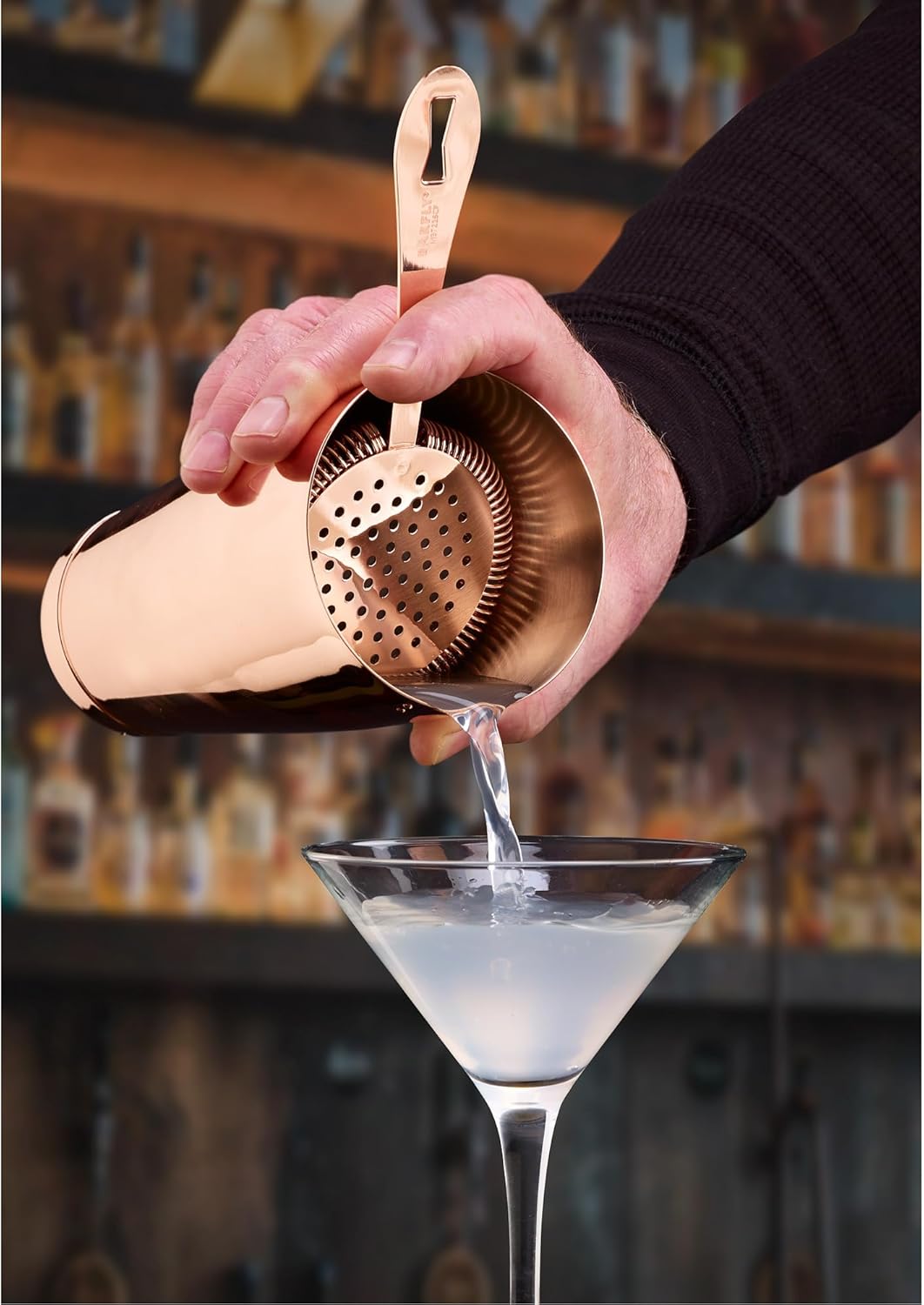 A bartender pouring a cocktail from a shaker with a Copper plated Barfly Deluxe Antique Hawthorne Spring Strainer on bar setting as a background