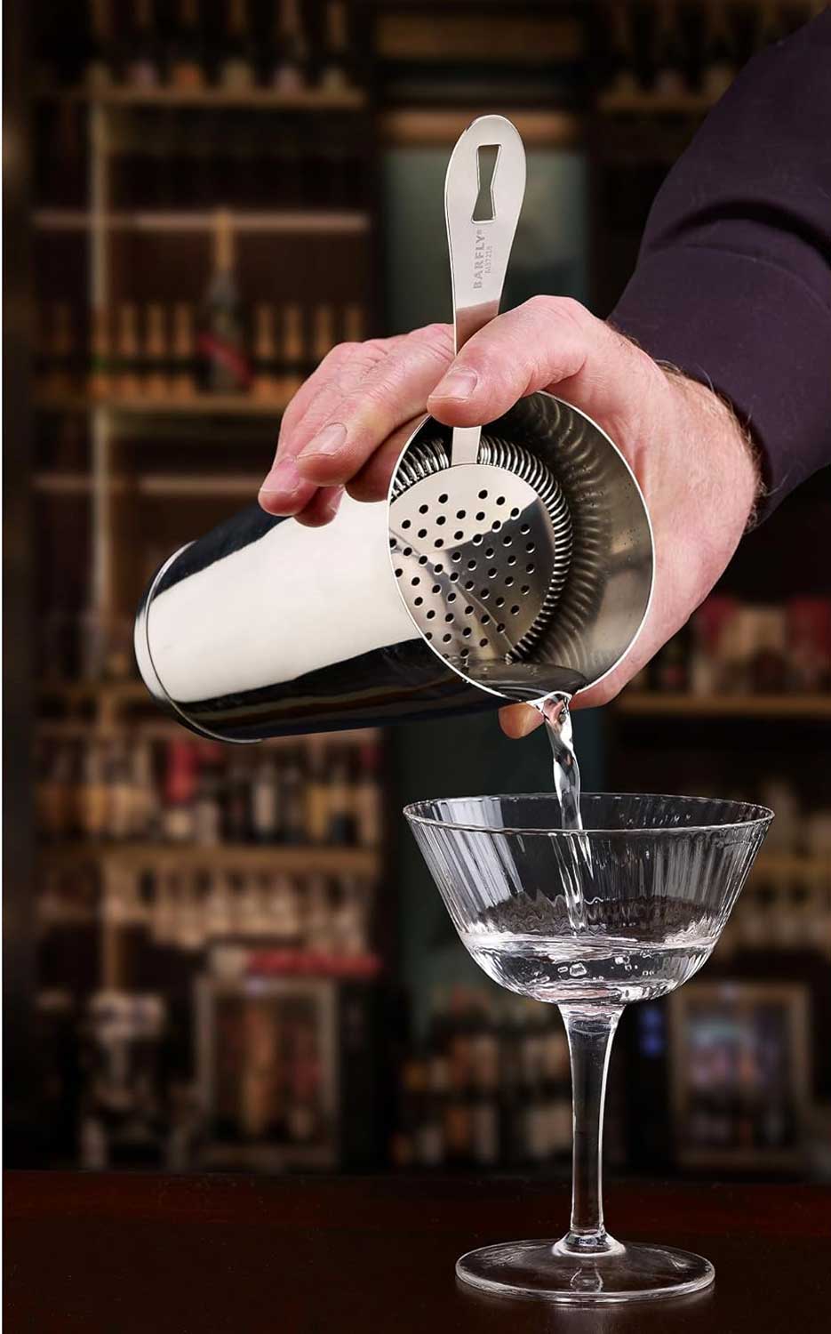 A bartender pouring a cocktail from a shaker with a stainless steel Barfly Deluxe Antique Hawthorne Spring Strainer into a glass with a blurred bar background