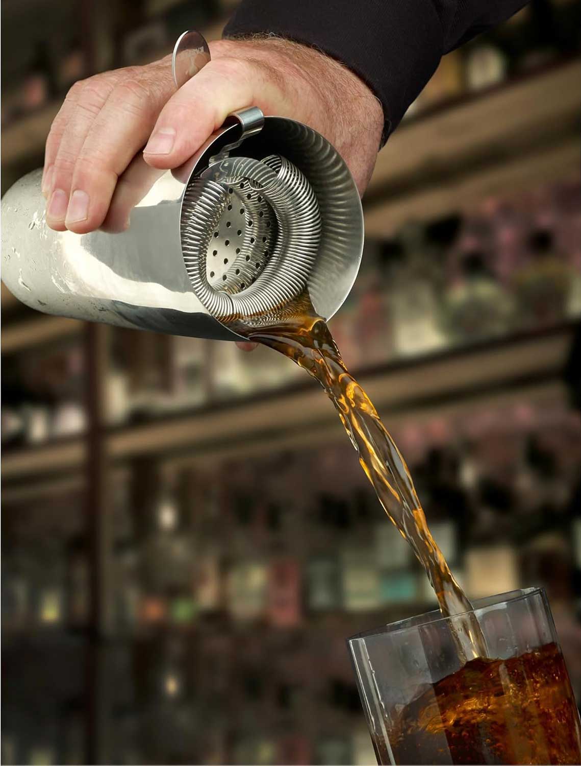 A bartender pouring a cocktail from a shaker with a Stainless Steel Barfly Throwing Strainer on a glass with ice
