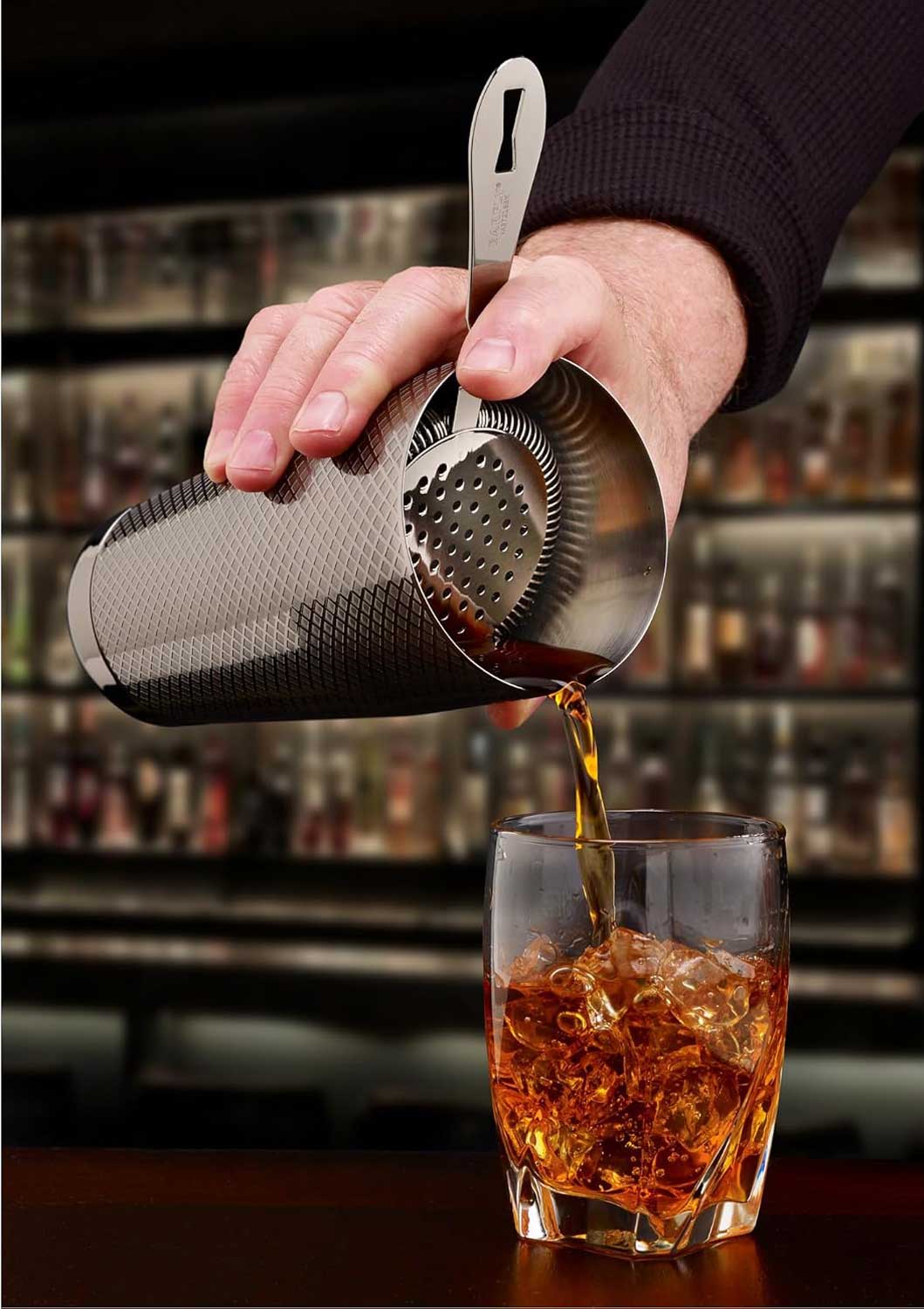 A bartender pouring a cocktail from a shaker with a Gun Metal Black colored Deluxe Antique Hawthorne Spring Strainer on a glass with ice