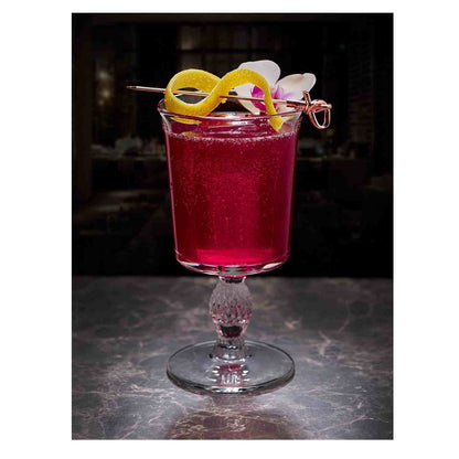 A deep magenta cocktail in a stemmed glass with a decorative, diamond-cut base. The drink is garnished with a lemon twist and a white orchid flower, skewered on a 4.5 inch Copper Plated Barfly Cocktail Pick, with a bamboo knot top.
