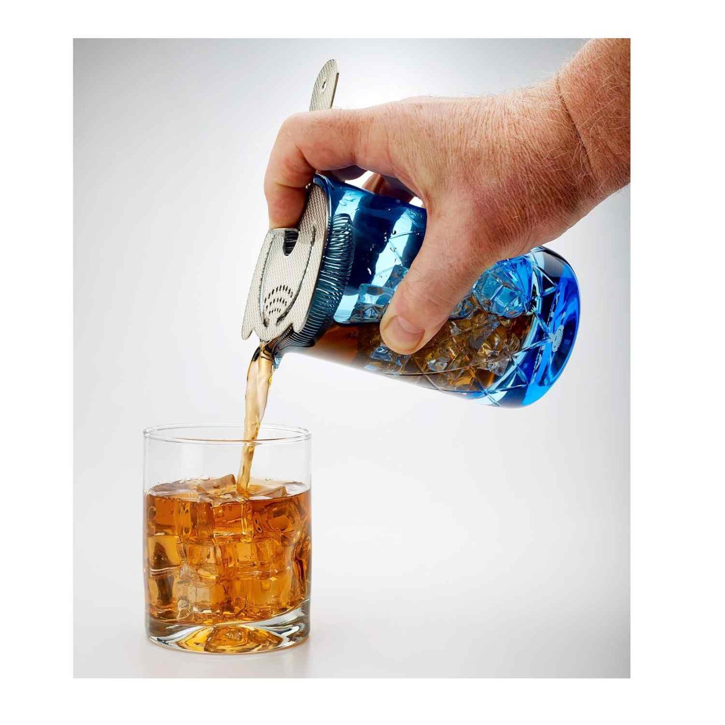 Hand pouring a cocktail from a close-up of the Blue Barfly Mixing Glass 17oz (500 ml)  with a bar strainer into a glass with ice cubes on a white background