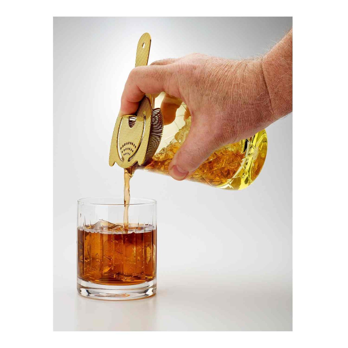 Hand pouring a cocktail from a close-up of the Gold Barfly Mixing Glass 17oz (500 ml)  with a bar strainer into a glass with ice cubes on a white background