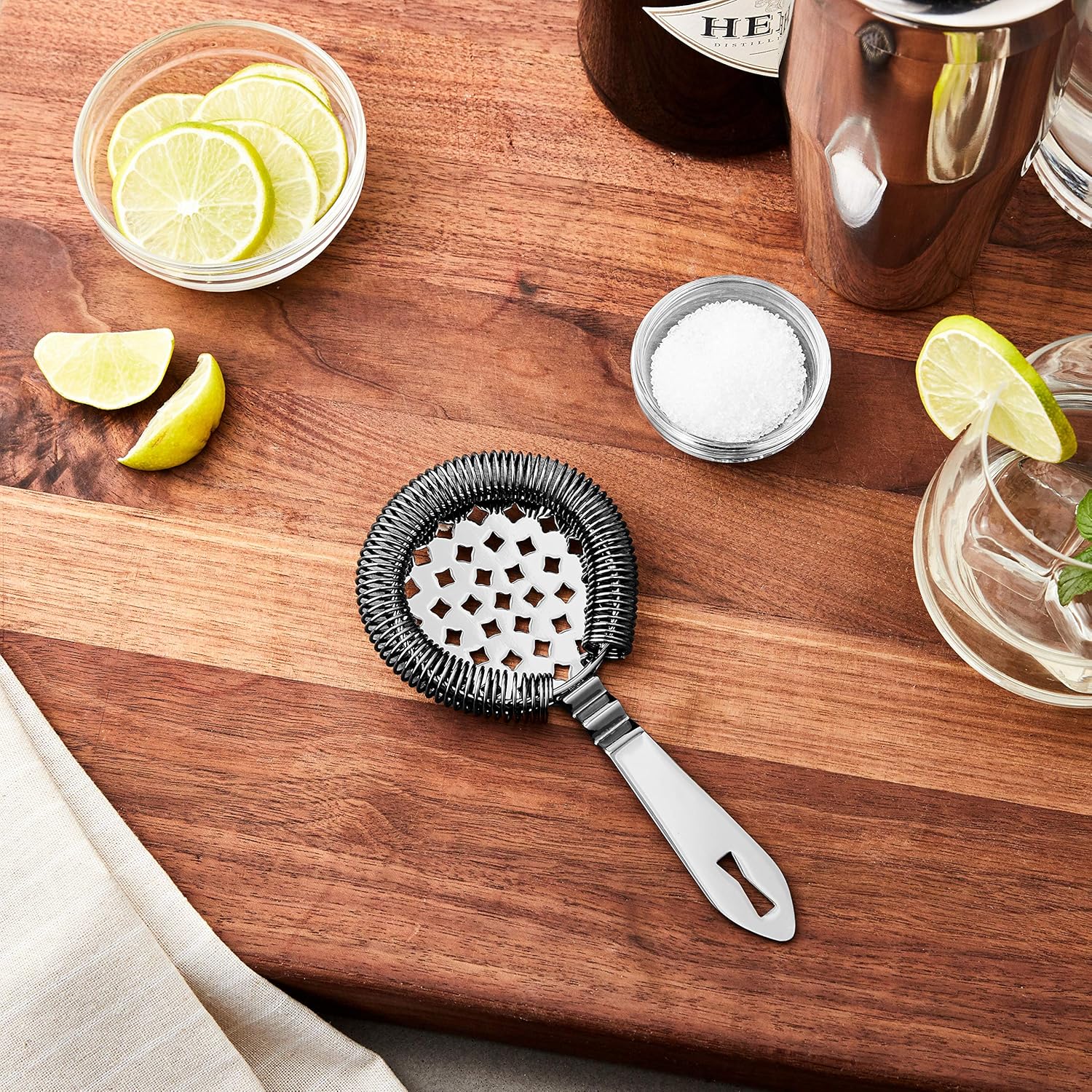 A Gun Metal Black colored Barfly Classic Hawthorne Spring Bar Strainer on a wooden surface with limes, salt, and a cocktail glass.