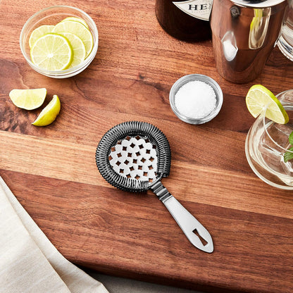 A Gun Metal Black colored Barfly Classic Hawthorne Spring Bar Strainer on a wooden surface with limes, salt, and a cocktail glass.