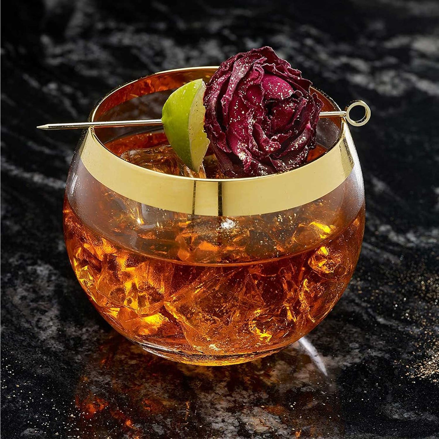 A golden-rimmed glass holding an amber cocktail filled with crushed ice. The drink is garnished with a 4.5 inch Gold Plated Barfly Cocktail Pick, with a Circle top, skewering a dried red rose and a lime wedge.