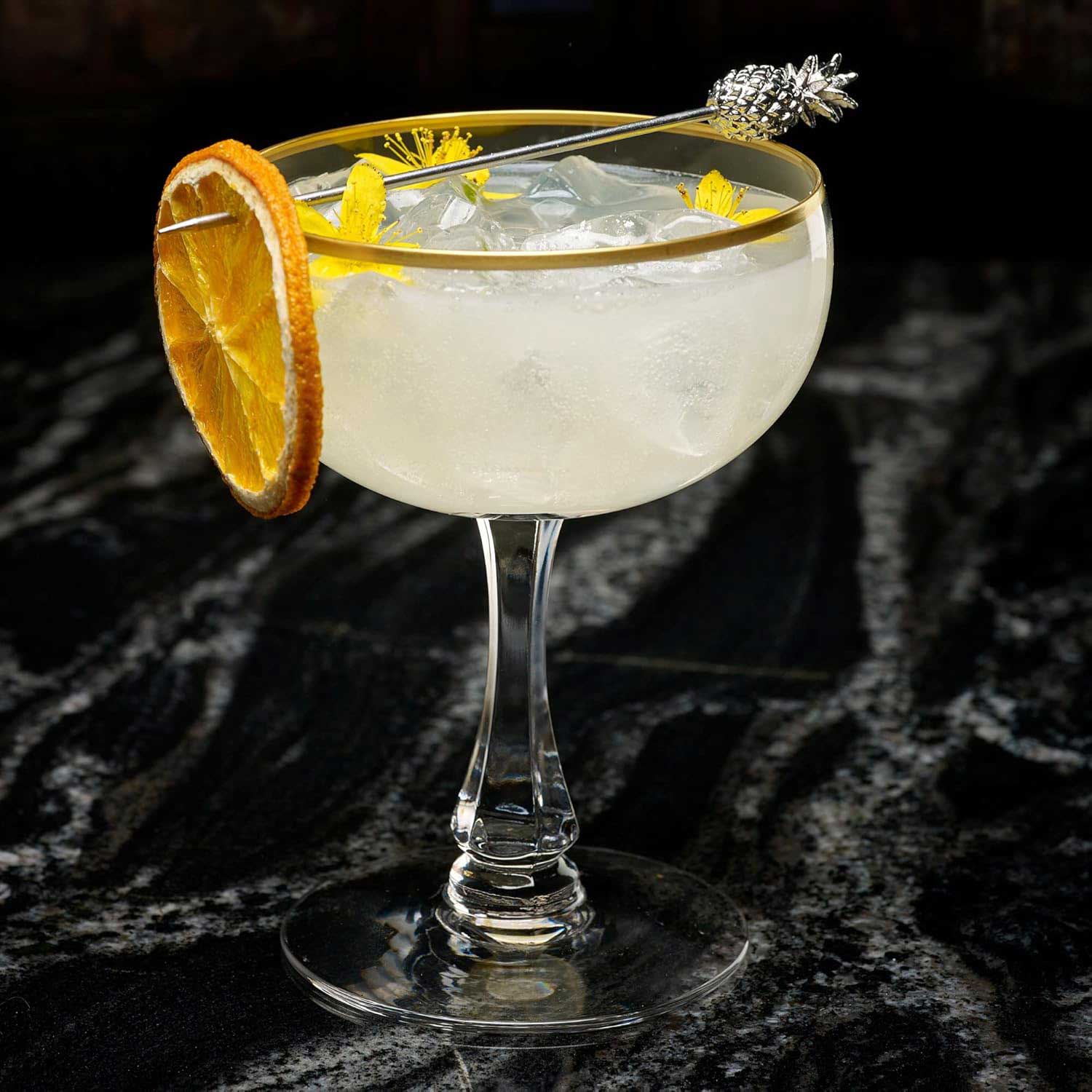 Cocktail in a coupe glass with a 4.5 inch Stainless Steel Barfly Cocktail Pick, with a Pineapple on the end and orange wheel on a dark background