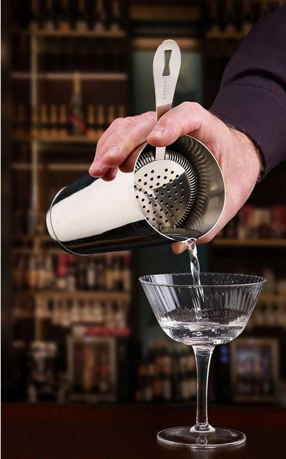 A bartender pouring a cocktail from a shaker with a stainless steel Barfly Deluxe Antique Hawthorne Spring Strainer into a glass with a blurred bar background