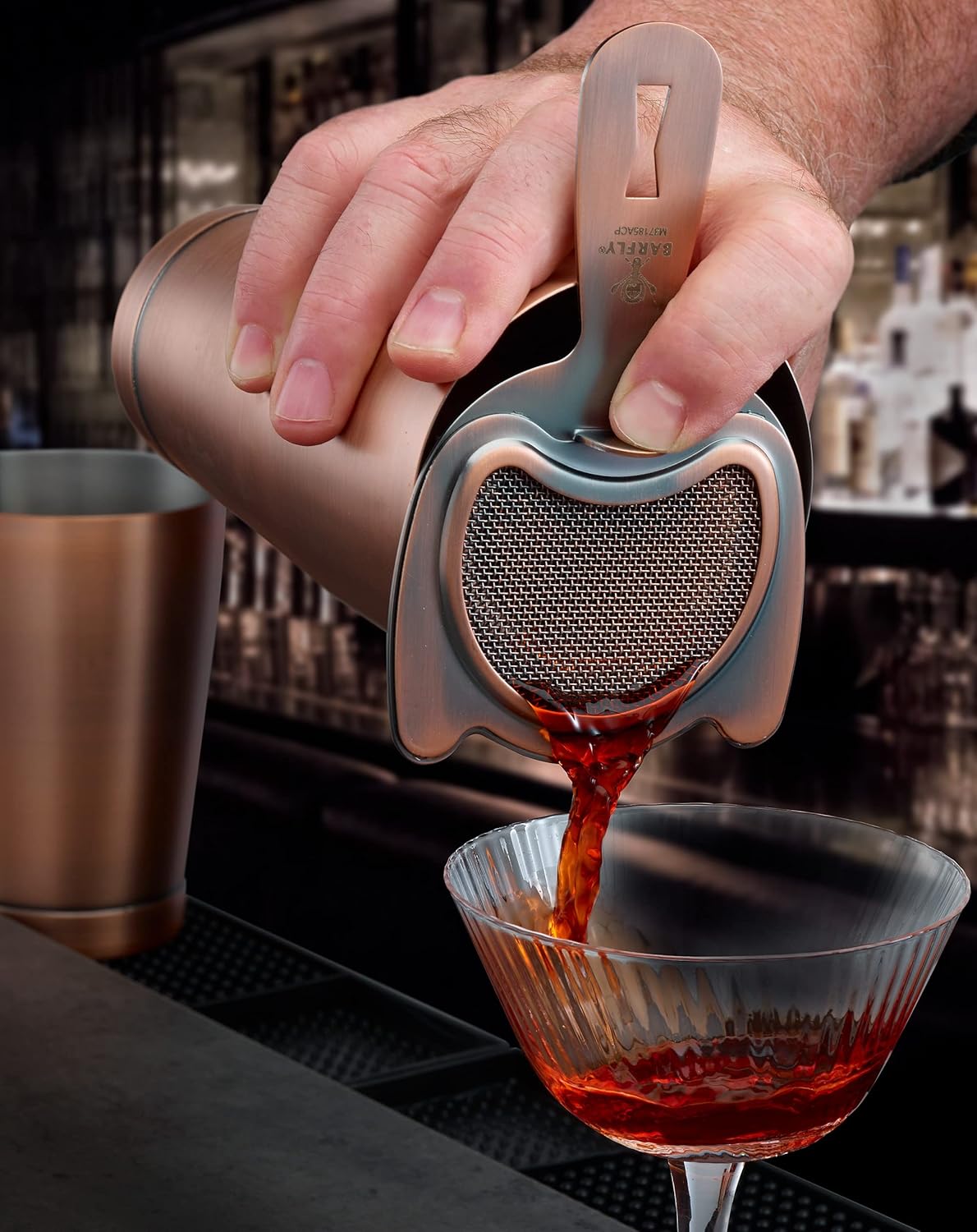 A bartender pouring a cocktail from a shaker with an Antique Copper finished Barfly Fine Mesh Spring Strainer on a glass
