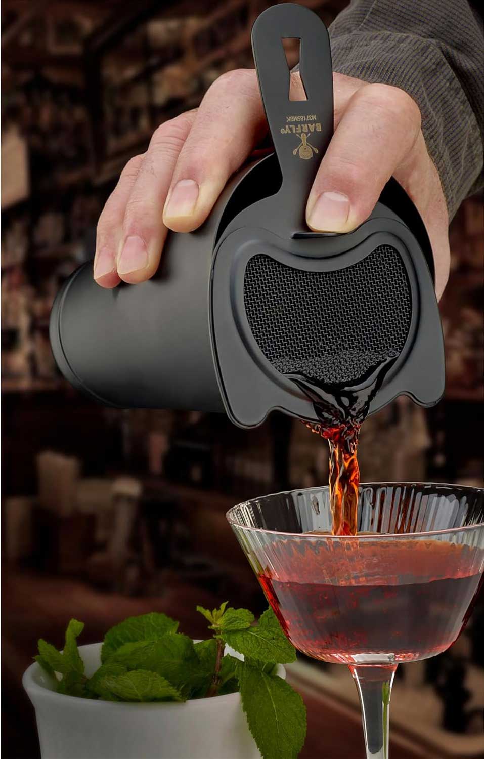A bartender pouring a cocktail from a shaker with a Matte Black Barfly Heavy-Duty 4 Prong Spring Bar Strainer into a glass with a blurred bar background
