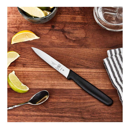 A 4 inch black Mercer Culinary Pointed Tip Wavy Bar Knife on a wooden surface with lemon and lime wedges, a spoon, and a striped towel.