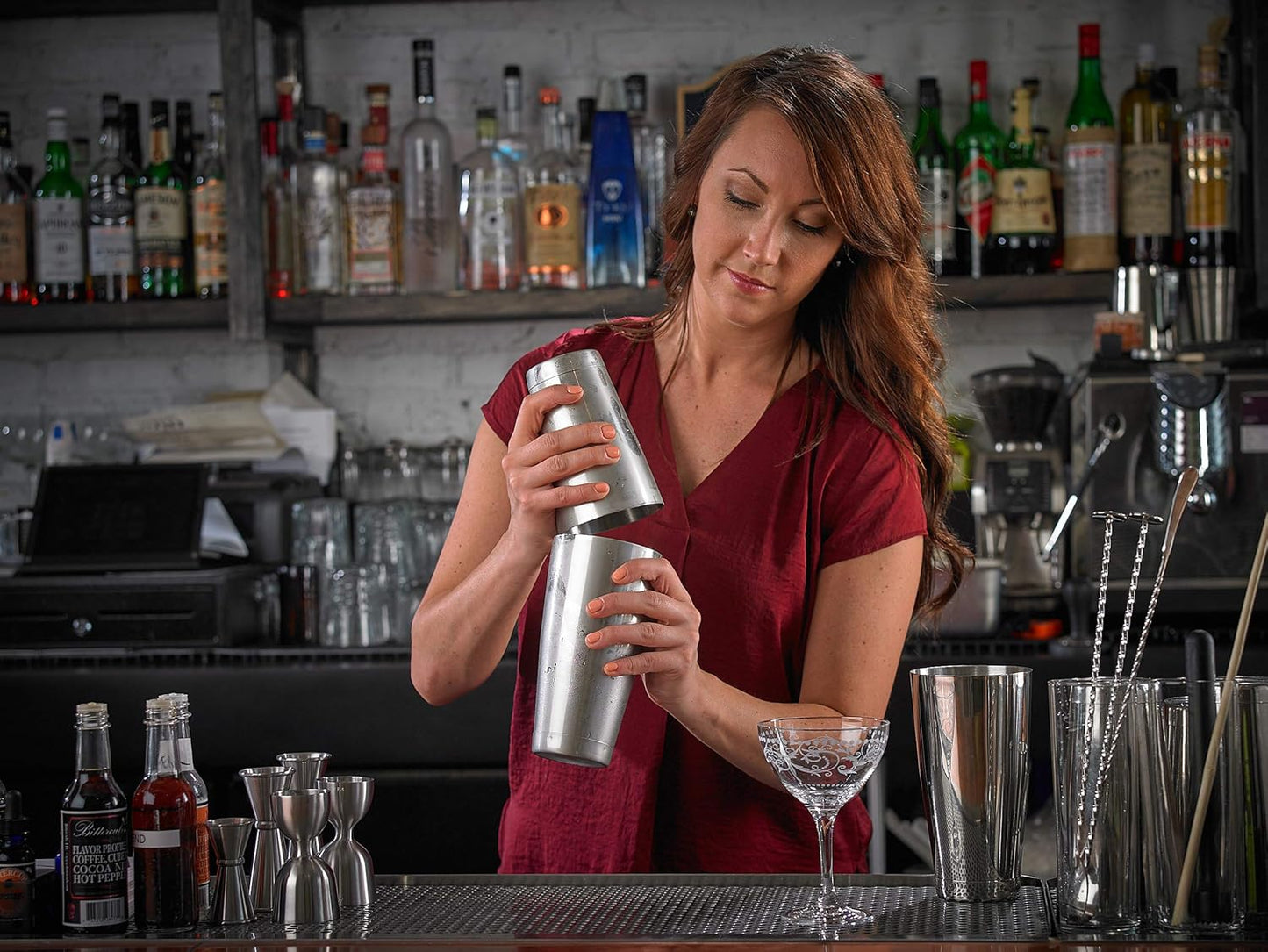 Woman in a red shirt preparing a cocktail using a barfly stainless steel shaker cocktail tin set of 18 oz and 28 oz in a bar setting with various bottles and glasses.
