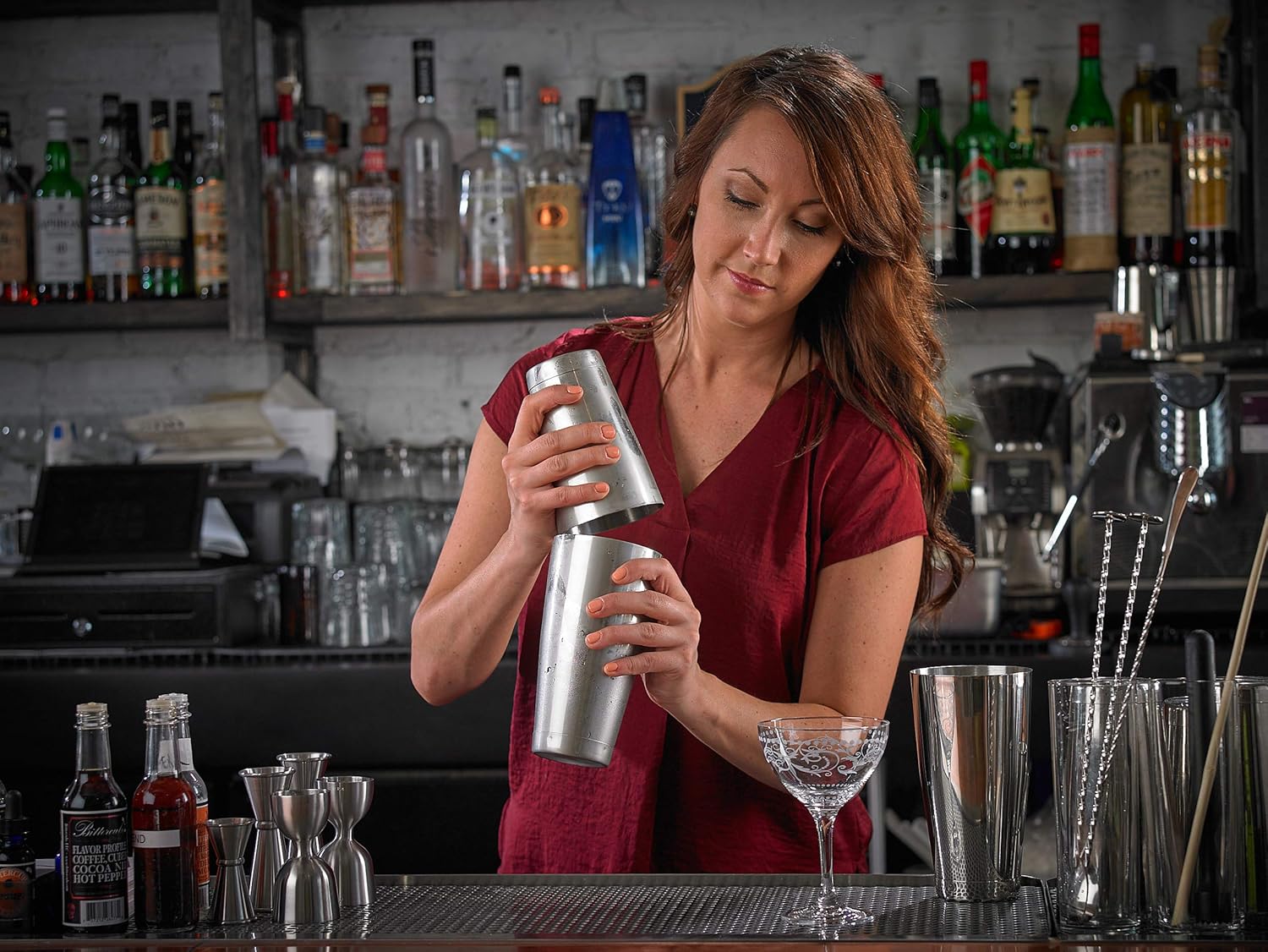 Woman in a red shirt preparing a cocktail using a barfly stainless steel shaker cocktail tin set of 18 oz and 28 oz in a bar setting with various bottles and glasses.