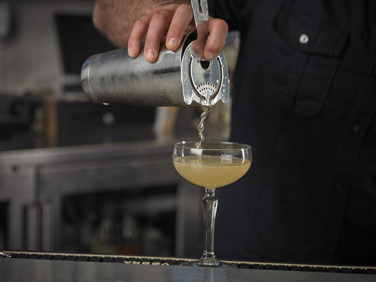Person pouring a cocktail from a barfly stainless steel shaker cocktail tin set of 18 oz and 28 oz into a glass on a bar.