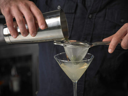 Person pouring a cocktail from a barfly stainless steel shaker cocktail tin set of 18 oz and 28 oz into a martini glass using a strainer.
